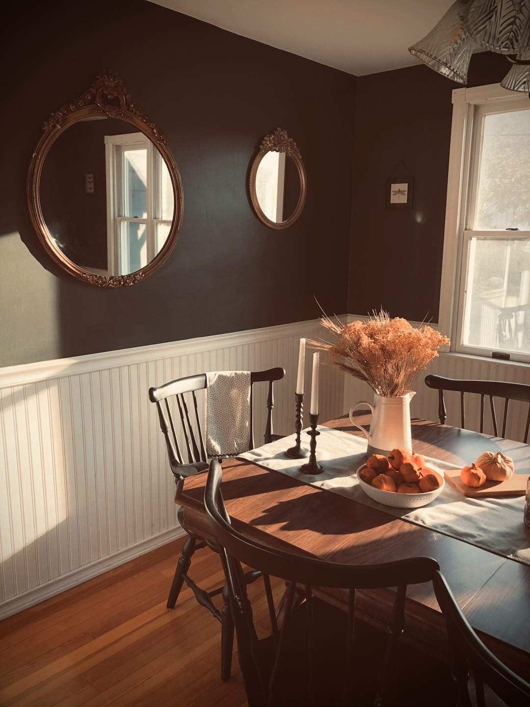 Antique dark brown wooden table surrounded by vintage black chairs in a dark green walled dining room. Ornate vintage gold mirrors hang on the walls. Faux pumpkins and dried foliage decorate the tabletop along with two black candlestick holders.
